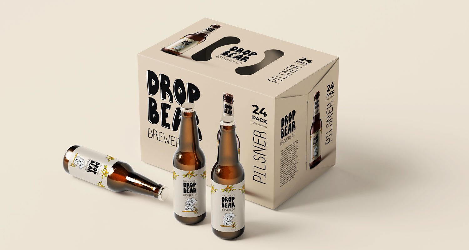 Photograph of a 24-case of beer featuring the Drop Bear brand identity. In front of the case are three bottles with labels showcasing an illustration of three cute koalas stacked on a tree branch, using Australian-inspired colors. The case and bottles are isolated against a neutral background.