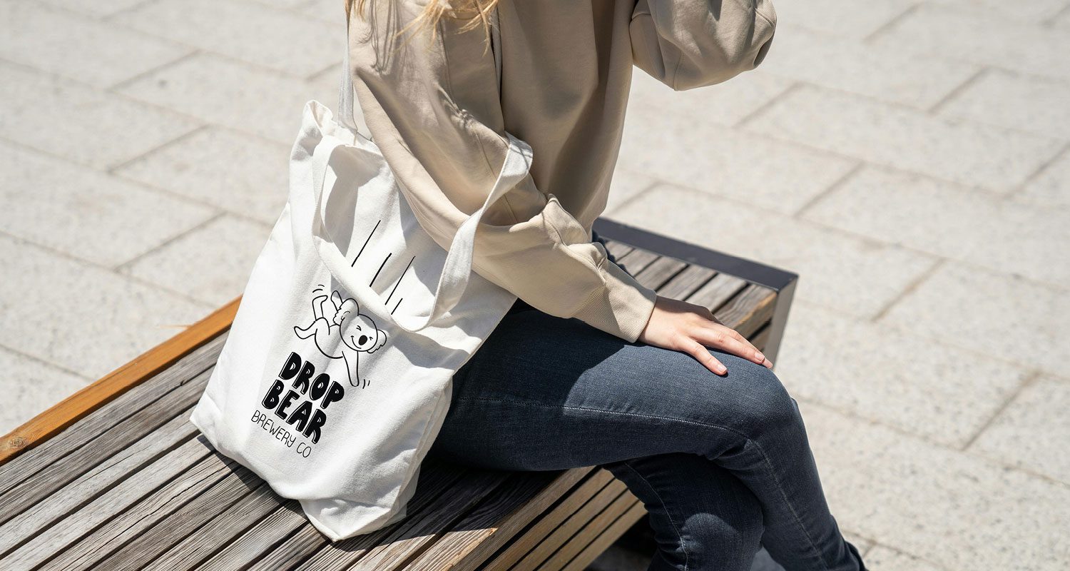 Photograph of a woman sitting on a street bench, with a tote bag resting on the wooden seat. The tote features a custom illustration of a cheeky koala falling, with the Drop Bear logo design placed below the koala.