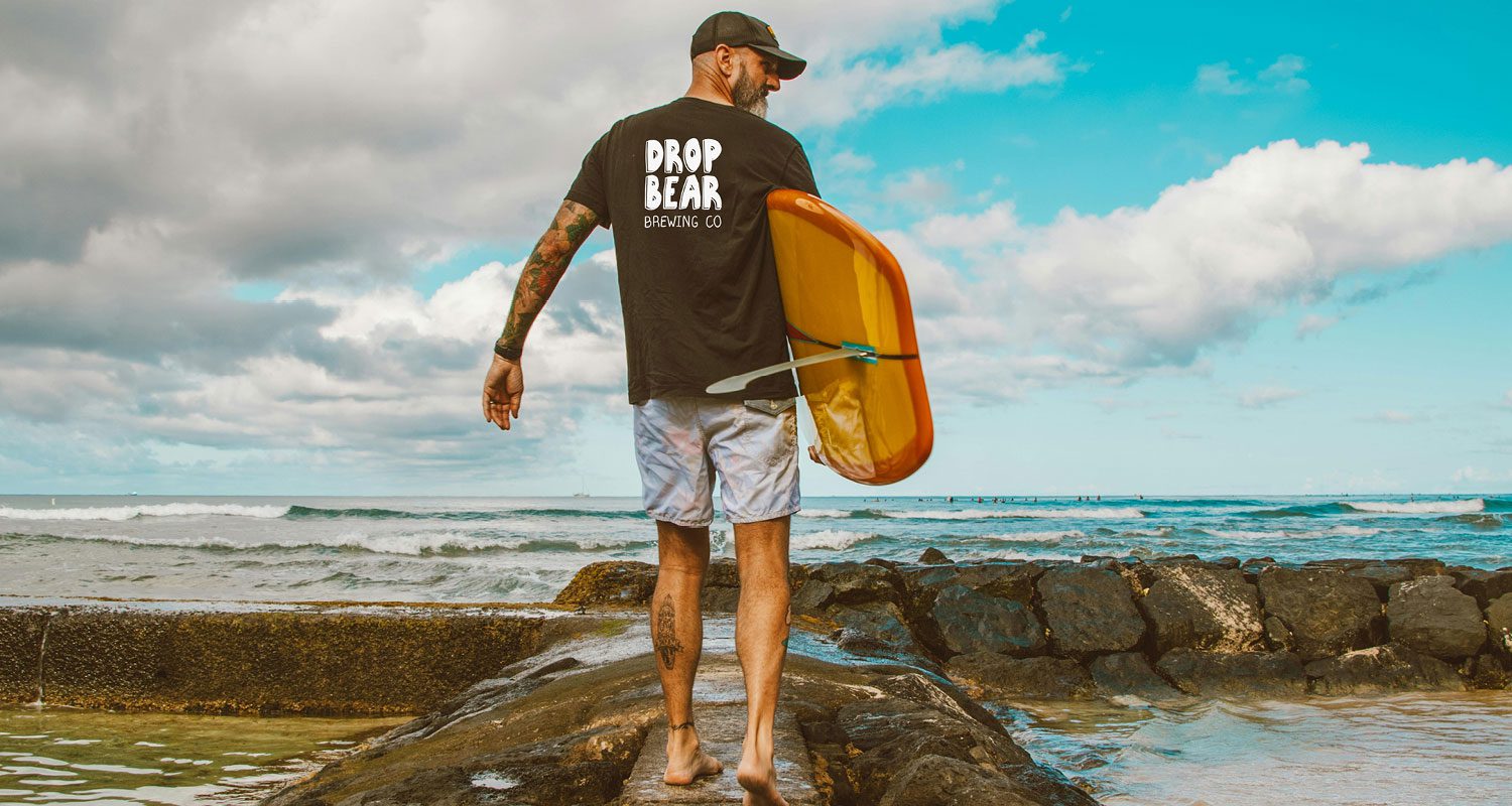 Photograph of a man walking along rocks toward the ocean beach, carrying a surfboard. The back of his black t-shirt prominently displays the Drop Bear brand logo design.