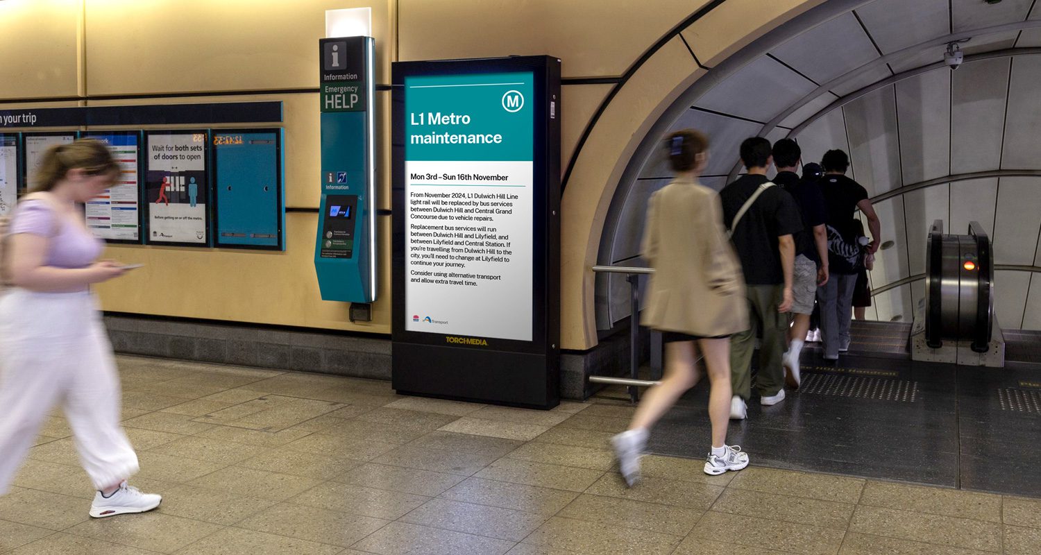 Photograph of a Transport for NSW operational template displayed on a digital banner at a Sydney Metro station, with a variety of people walking past to catch the Metro.