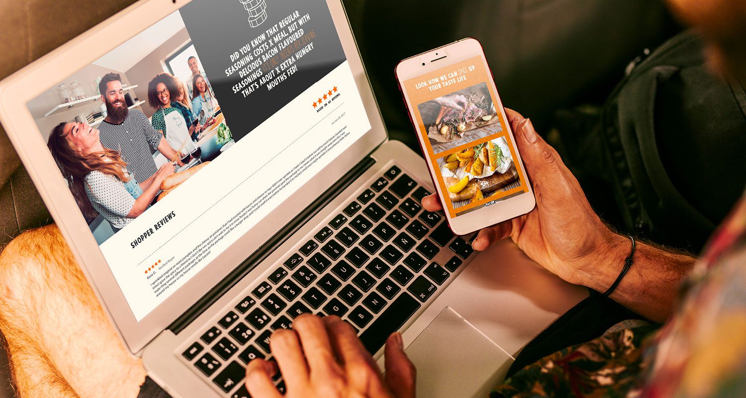 Photograph of a person holding a mobile phone in one hand while working on a computer, both screens displaying the Deliciou Seasoning web design, showcasing a seamless digital experience across devices.