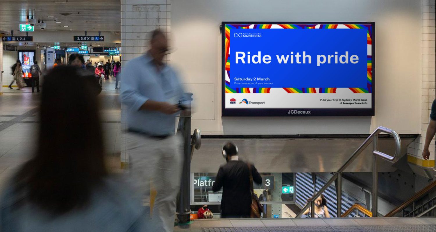 A vibrant photograph taken inside a train station, showcasing a pride-themed digital screen that reads “Ride with Pride.” The scene features a diverse group of passengers—some walking, others waiting—set against a colorful, well-lit train station interior. The campaign, led by Transport for NSW, celebrates LGBTQ+ inclusion and visibility in public transport spaces.