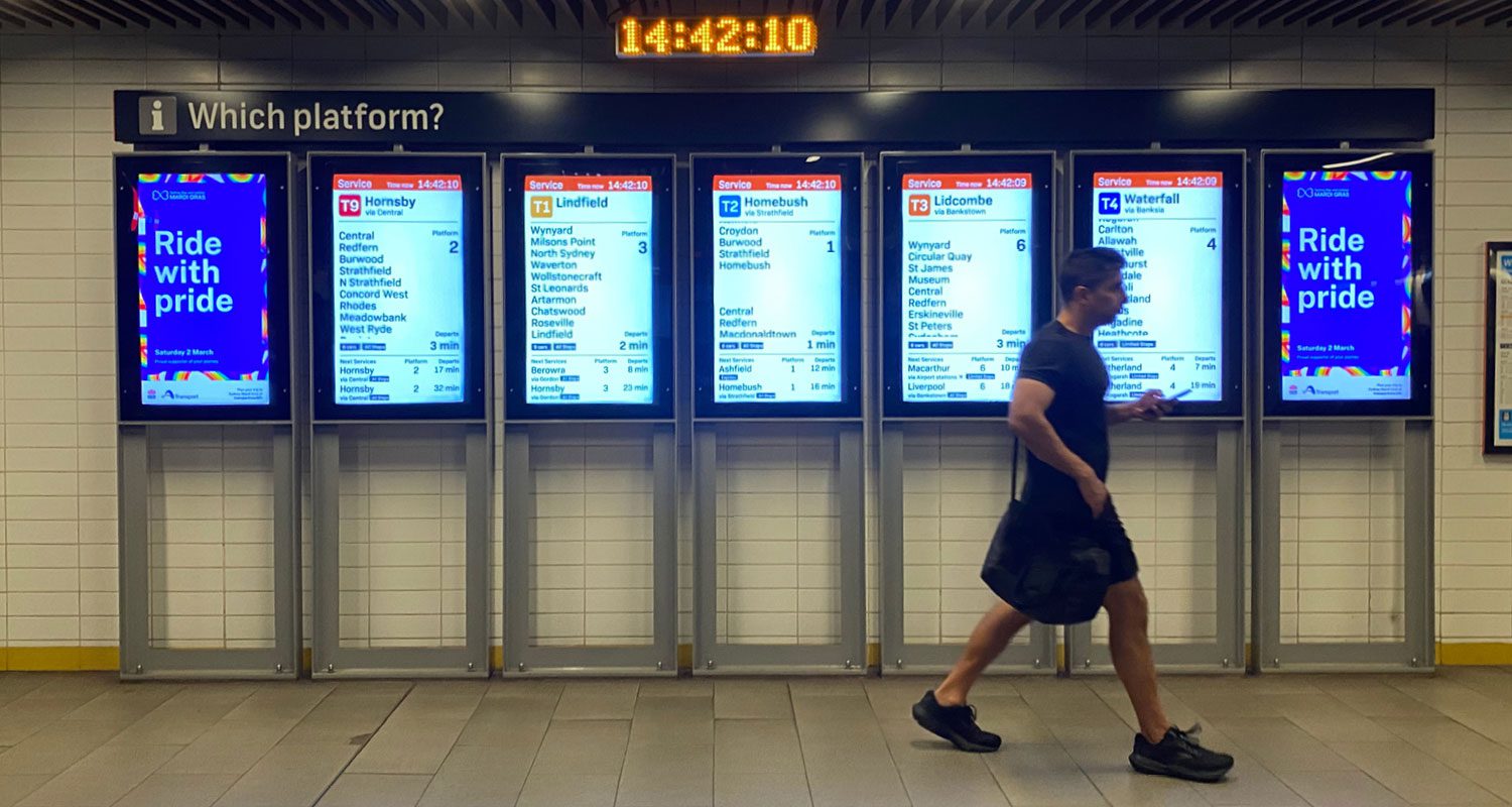 Photograph of a pride-themed Transport for NSW poster displayed on digital screens inside a train station, with a person walking past, featuring the “Ride with Pride” message and a colorful rainbow design.