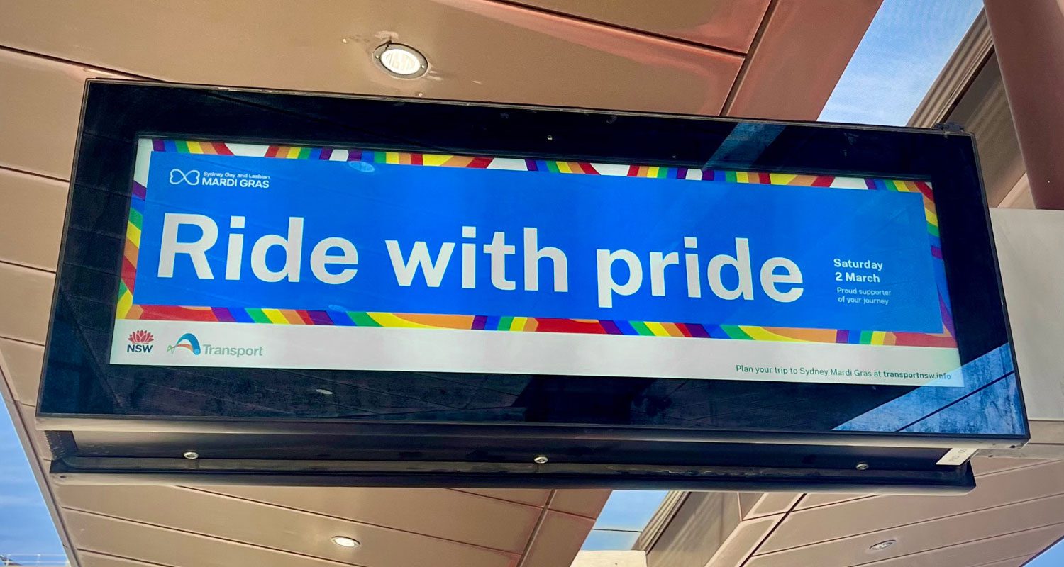 Photograph of a colorful public transport scene featuring a wide digital screen at a light rail station backdrop, promoting inclusivity and the “Ride with Pride” campaign by Transport for NSW.